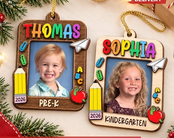 May include: Two rectangular wooden photo frame ornaments with gold hanging strings. One frame is dark wood with the name THOMAS and PRE-K, the other is light wood with SOPHIA and KINDERGARTEN. Both feature colorful pencil, paper plane, and apple icons.
