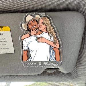 May include: A clear acrylic car visor clip featuring a cartoon illustration of a person in a cowboy hat being hugged from behind by another person with long blonde hair. The text Forever & Always is printed in white at the bottom of the ornament.