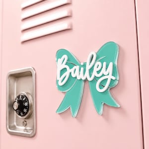 May include: A teal acrylic bow-shaped sign featuring the name Bailey in white script lettering. The sign is attached to a light pink metal locker door with a silver combination lock visible to the left of the bow-shaped name plate.
