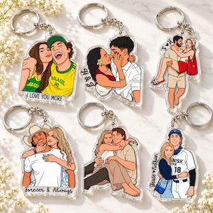 May include: Six clear acrylic keychains featuring illustrated couples in various poses and outfits. Designs include a Brazil soccer jersey, a couple embracing, a person carrying another, a cowboy hat, a seated pair, and a baseball uniform with text.