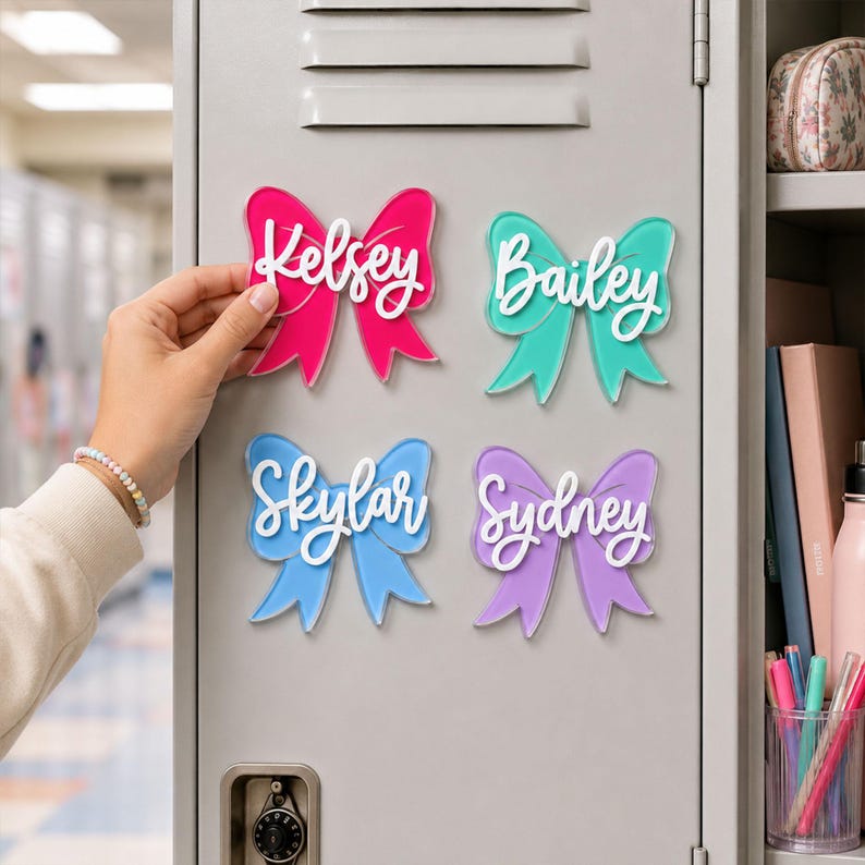 May include: Four bow-shaped acrylic magnets attached to a gray metal locker. Each magnet features a different color&mdash;hot pink, teal, light blue, and lavender&mdash;with a white script name layered on top: Kelsey, Bailey, Skylar, and Sydney.