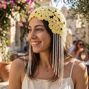 May include: A pale yellow crochet cap featuring a floral pattern with long, dangling strands of white faux pearls hanging from the edges. The headpiece is constructed with intricate yarn loops and delicate beadwork that frames the face.