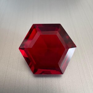 May include: A faceted, hexagonal gemstone in a deep, rich red hue. The gem's multiple facets catch the light, creating a brilliant display. The shape is symmetrical, with sharp angles and a polished surface. The gemstone is set against a neutral background.