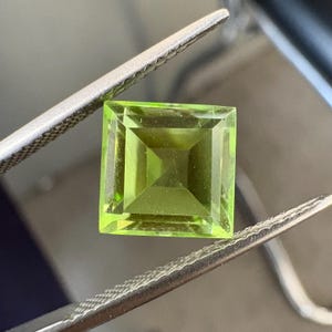 May include: A square-cut, translucent lime green gemstone with a faceted surface. The stone is held by a pair of silver-toned metal tweezers, showcasing its sharp edges and geometric shape. The gem displays a vibrant, light-reflecting quality.