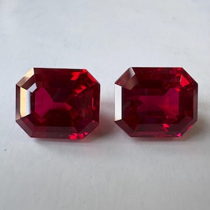 Può includere: Due pietre preziose rettangolari a taglio smeraldo di un colore rosso profondo e vibrante. Ogni pietra presenta superfici sfaccettate che riflettono la luce, creando un aspetto chiaro e trasparente con bordi netti e definiti.