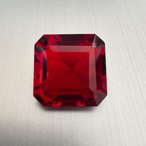 May include: A faceted, rectangular-cut, deep red gemstone. The gem has a square shape with beveled edges, reflecting light and showcasing its rich color. The gemstone is set against a light background.