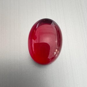 Könnte beinhalten: Ein polierter, oval geformter, tiefroter Edelstein. Der Stein hat eine glatte, reflektierende Oberfläche mit einem Farbverlauf von einem dunkleren Farbton oben zu einem helleren Rot an der Basis. Der Edelstein ist vor einem hellgrauen Hintergrund platziert.