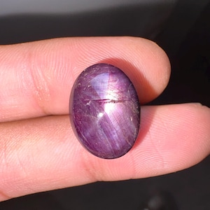 May include: An oval-shaped cabochon star ruby gemstone with a deep purple-red hue. The stone features a distinct six-rayed star effect, known as asterism, visible on its smooth, polished surface, reflecting light in a bright, centered pattern.