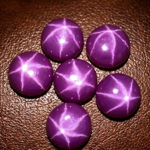 May include: Six round, cabochon-cut purple gemstones arranged in a cluster. Each stone features a distinct six-rayed star pattern, known as asterism, centred on the smooth, polished surface, reflecting light with a bright, white, star-shaped glow.