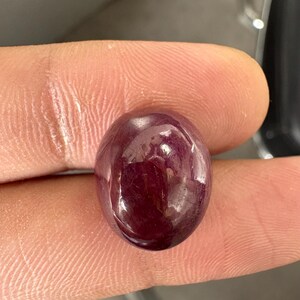 May include: A deep, dark red gemstone with a smooth, polished cabochon cut. The stone features a rounded, oval shape with a glossy, reflective surface that displays subtle internal inclusions and variations in its rich, saturated crimson color.