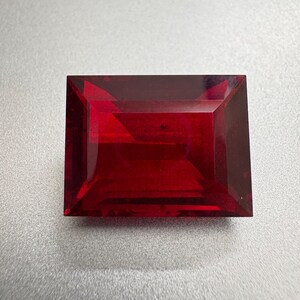 Puede incluir: Una gema rectangular facetada de color rojo intenso. La gema tiene un corte geométrico limpio, que muestra su claridad y brillo. Las facetas capturan la luz, creando un juego de sombras y reflejos. La gema se encuentra sobre un fondo neutro.