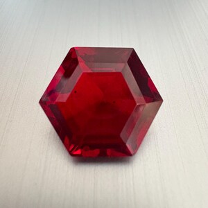 May include: A faceted, hexagonal-shaped, deep red gemstone. The gem has sharp angles and a polished surface, reflecting light. The overall shape is symmetrical, with a flat top and a slightly translucent appearance.