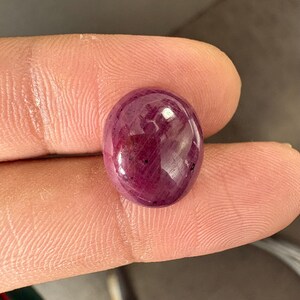 Puede incluir: Una piedra preciosa tipo cabujón de forma ovalada y pulida con un color rojo magenta profundo y saturado. La piedra presenta una superficie lisa y redondeada con inclusiones internas visibles y variaciones naturales en su tono translúcido.