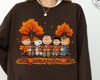May include: A dark brown crewneck sweatshirt featuring a graphic print of the Peanuts characters sitting on a wooden bench under autumn trees, each holding a mug. The design includes orange fall leaves and pumpkins on the ground.