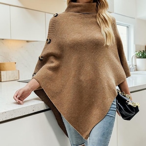 Peut inclure: Un poncho marron avec un col roulé et des boutons sur l'épaule. Le pull est en maille douce et a une coupe ample. Le modèle porte un jean bleu et tient un sac à main noir.