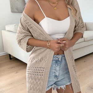 Puede incluir: Cárdigan beige con frente abierto y un patrón de punto geométrico. El cárdigan tiene mangas tres cuartos y se usa sobre una camiseta blanca y pantalones cortos de mezclilla. Collares y pulseras doradas completan el look.
