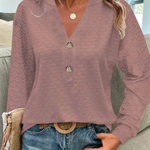 Pode incluir: Uma blusa de manga comprida cor malva com decote em V e dois botões decorativos. A blusa tem um delicado padrão vazado e é combinada com jeans azuis e um cinto castanho com fivela circular. Um colar de ouro é visível.