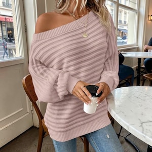 May include: A pale pink off-the-shoulder jumper with long sleeves. The jumper features a ribbed knit pattern and is worn with blue jeans. A gold necklace and a coffee cup with a black lid are also visible.