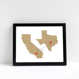 May include: Framed art print featuring wooden-textured outlines of California and Texas, connected by a dotted line with red heart markers. The artwork is set against a white background, framed in a black border.