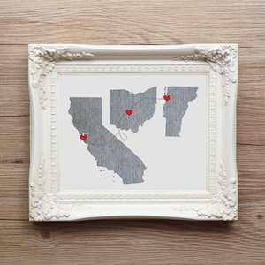 May include: A white framed print with a map of the United States showing a dotted line connecting California, Ohio, and Maine. Red hearts mark the locations on the map.