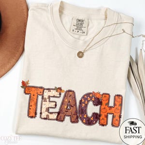 May include: A cream-colored short-sleeve t-shirt with a Comfort Colors label at the neck. The front features the word TEACH in large block letters, each filled with different autumn and Halloween patterns including ghosts, pumpkins, and candy corn.