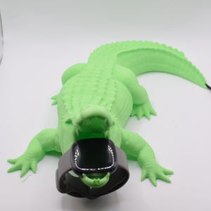 May include: A light green alligator-shaped charging stand holding a smartwatch. The alligator has a textured surface and is lying on a white surface. A black charging cable extends from the alligator's tail.