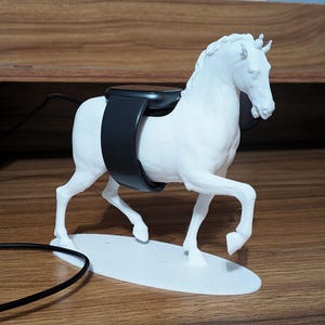 May include: A white horse-shaped charging stand for a smartwatch. The black smartwatch band is wrapped around the horse's back. The stand is on a white oval base, with a black charging cable attached.