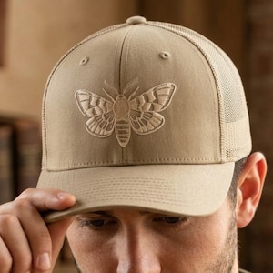 May include: Tan baseball cap with a mesh back and a moth design embroidered on the front. The cap has a curved brim and a structured crown. The moth design is in a lighter tan color.