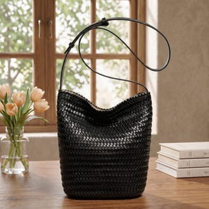 Op de afbeelding: Een zwarte geweven leren emmer tas met een enkele schouderriem. De tas heeft een cilindrische vorm met een brede opening en een geweven patroon. De tas staat op een houten tafel. Links een boeket tulpen in een glazen vaas.