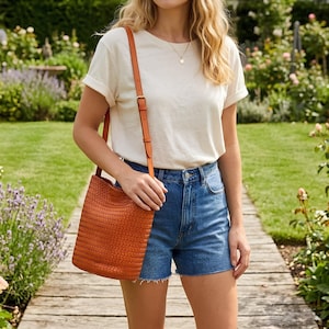 Op de afbeelding: Een geweven leren emmer tas in een warme bruine tint, met een schouderriem en een gesp. De tas wordt vastgehouden door een persoon die een wit t-shirt en een spijkerbroek draagt. De achtergrond toont een tuin met een houten pad.