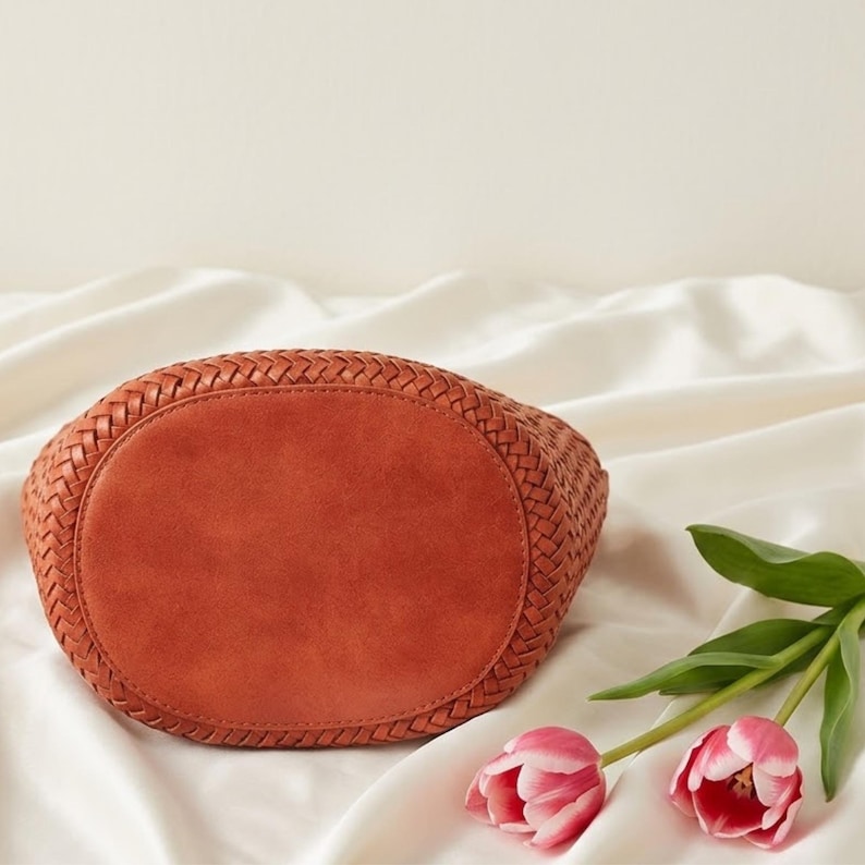 Op de afbeelding: Een geweven, roestkleurige handtas met een afgeronde basis. De bodem van de tas is ovaal. Twee roze tulpen met groene stelen en bladeren liggen op de voorgrond, op een witte, zijdeachtige ondergrond.