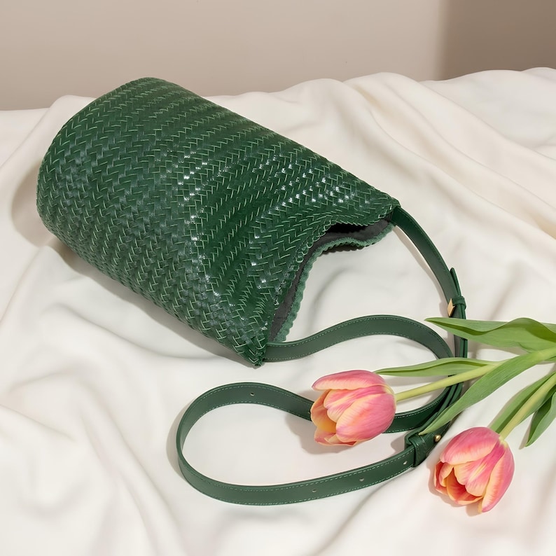 Op de afbeelding: Een groene geweven handtas met een schouderriem. De tas is cilindrisch met een brede opening. Twee roze en gele tulpen liggen op een witte doek naast de tas.