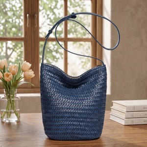 Op de afbeelding: Een marineblauwe geweven leren emmer tas met een lange schouderriem. De tas staat op een houten tafel met een vaas met oranje tulpen en een stapel boeken op de achtergrond. De tas heeft een uniek, open ontwerp.