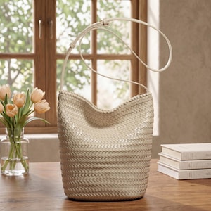 Op de afbeelding: Een beige geweven emmertas met een lange, dunne riem. De tas heeft een afgeronde vorm en een textuur. De riem is met een simpele knoop aan de tas bevestigd. De tas staat op een houten oppervlak.