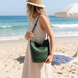 Op de afbeelding: Een donkergroene, geweven leren schoudertas met een lange riem. De tas wordt vastgehouden door een persoon op een zandstrand met de oceaan op de achtergrond. Een strohoed en een parasol zijn ook zichtbaar.