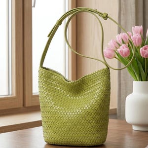 Op de afbeelding: Een geweven groene schoudertas met een lange riem. De tas heeft een emmervorm en is gemaakt van een textuurrijk materiaal. De riem heeft een bijpassende groene kleur. De tas staat op een houten oppervlak bij een raam.