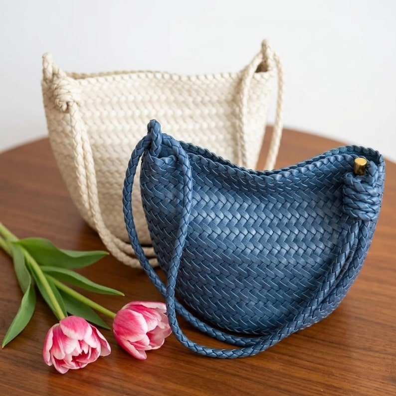 Op de afbeelding: Twee geweven handtassen in cr&egrave;me en blauw staan op een houten oppervlak, met roze en rode tulpen op de voorgrond. De blauwe tas heeft een gevlochten riem en een goudkleurige sluiting. De cr&egrave;mekleurige tas staat op de achtergrond.