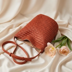 Op de afbeelding: Een geweven, roestkleurige handtas met een lange, verstelbare riem. De tas heeft een cilindrische vorm en een lichtgekleurde binnenkant. Twee roze bloemen met groene stelen liggen naast de tas op een witte satijnen ondergrond.
