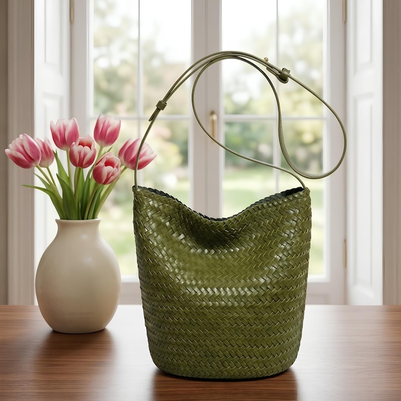 Op de afbeelding: Een olijfgroene geweven schoudertas met een lange riem. De tas is cilindrisch met een brede opening. Een vaas met roze en witte tulpen staat naast de tas op een houten tafel. De achtergrond toont een raam.