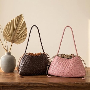 May include: Two woven handbags, one in dark brown and one in light pink, each featuring a braided shoulder strap, a drawstring closure, and a gathered interior lining. The bags have a structured, rectangular base and a textured, interlaced exterior.