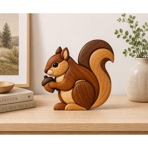 May include: A wooden squirrel figurine in shades of brown and tan, holding an acorn. The squirrel has a detailed, layered design and is posed in a sitting position. The figurine is on a wooden surface, with a framed picture and vase in the background.