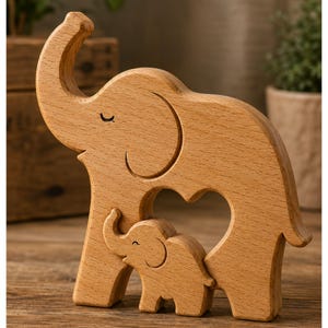 Op de afbeelding: Een lichtbruin houten beeldhouwwerk met een grote olifant met de slurf omhoog en een kleinere babyolifant geplaatst in een hartvormige uitsparing in het midden van het lichaam van de grote olifant, met zichtbare houtnerf.