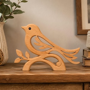 Peut inclure: Une figurine d'oiseau en bois brun clair présentant un design ajouré d'un oiseau perché sur une branche avec trois feuilles. L'oiseau a une finition lisse et un petit détail d'œil circulaire sombre, posé sur une base incurvée.