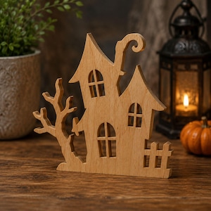 May include: A light brown wooden cutout of a two-story house with a pointed roof, a curved chimney, and arched windows. The structure features a bare tree on the left side and a small picket fence on the right side, all crafted from flat wood.