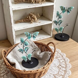 a wooden table with a woven basket containing a small plant with blue flowers, placed on a lace doily. In the background, there is a white shelf with dried flowers and a small sculpture of a butterfly.