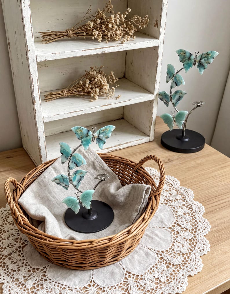 a wooden table with a woven basket containing a small plant with blue flowers, placed on a lace doily. In the background, there is a white shelf with dried flowers and a small sculpture of a butterfly.