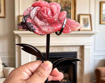 May include: A carved rhodonite stone rose sculpture featuring shades of pink, red, and black veining. The stone flower is mounted on a black metal stand with a thin stem, two pointed leaves, and a circular base with decorative cutouts.