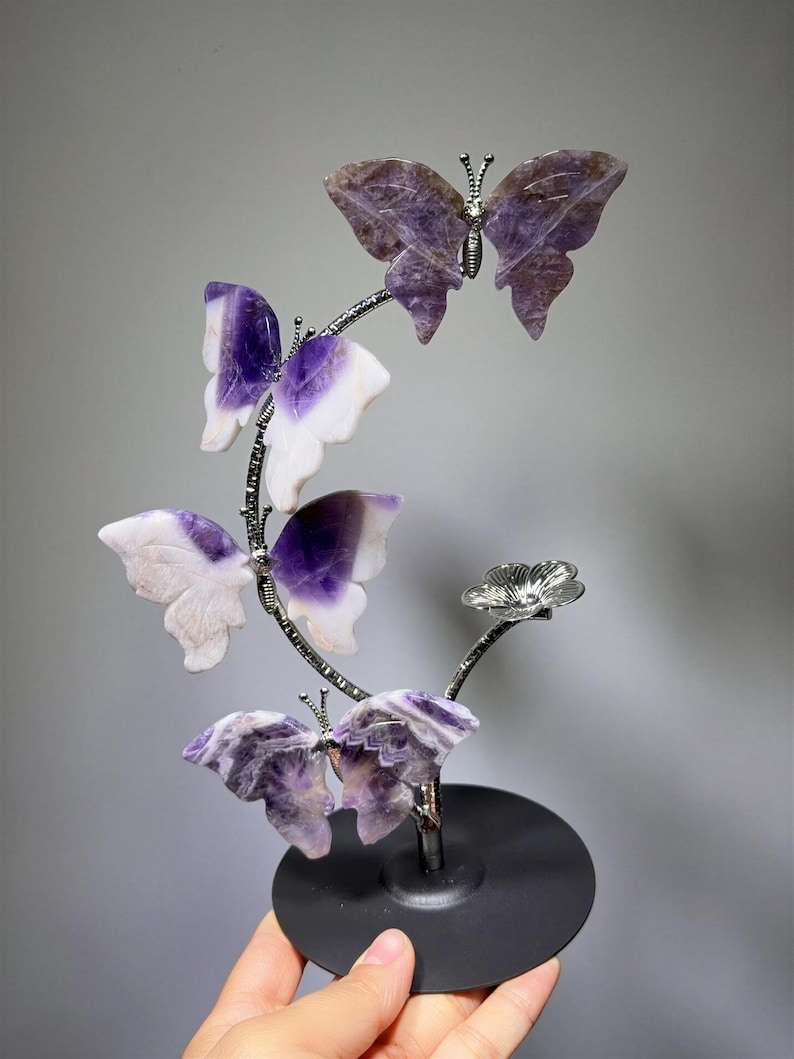 A hand is holding a decorative piece featuring a branch with purple and white butterfly-shaped ornaments.