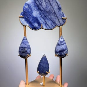 May include: A decorative sculpture featuring a blue, cloud-shaped stone piece suspended above three blue, teardrop-shaped stone pieces. The stones are held by a gold-toned metal frame consisting of a square base and vertical rods.