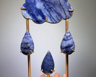 May include: A decorative sculpture featuring a blue, cloud-shaped stone piece suspended above three blue, teardrop-shaped stone pieces. The stones are held by a gold-toned metal frame consisting of a square base and vertical rods.